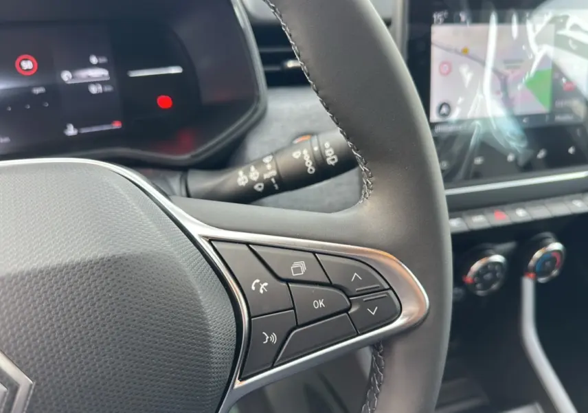 Gros plan sur le volant cuir multifonctions de la Renault Clio gris rafale, avec commandes intégrées et tableau de bord numérique en arrière-plan.