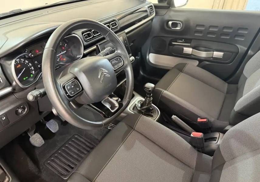 Vue intérieure côté conducteur de la Citroën C3 2019, tableau de bord noir avec volant cuir et sièges gris foncé.