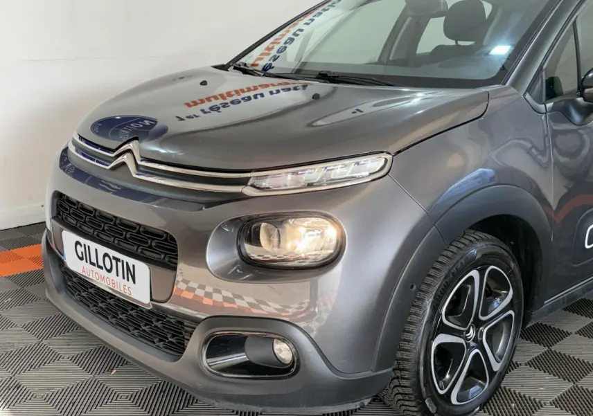 Vue 3/4 avant droit d'une Citroën C3 gris clair avec phares allumés et jantes alliage distinctives.