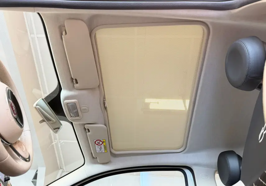 Vue intérieure du toit ouvrant avec pare-soleil beige de la Fiat 500 Dolcevita blanche, montrant le volant et appuie-tête noirs.