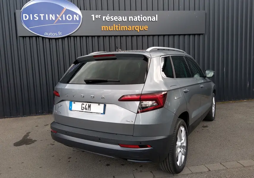 Vue 3/4 arrière droite d'un Skoda Karoq gris 2022 avec toit ouvrant et jantes alliage, stationné devant un mur noir.