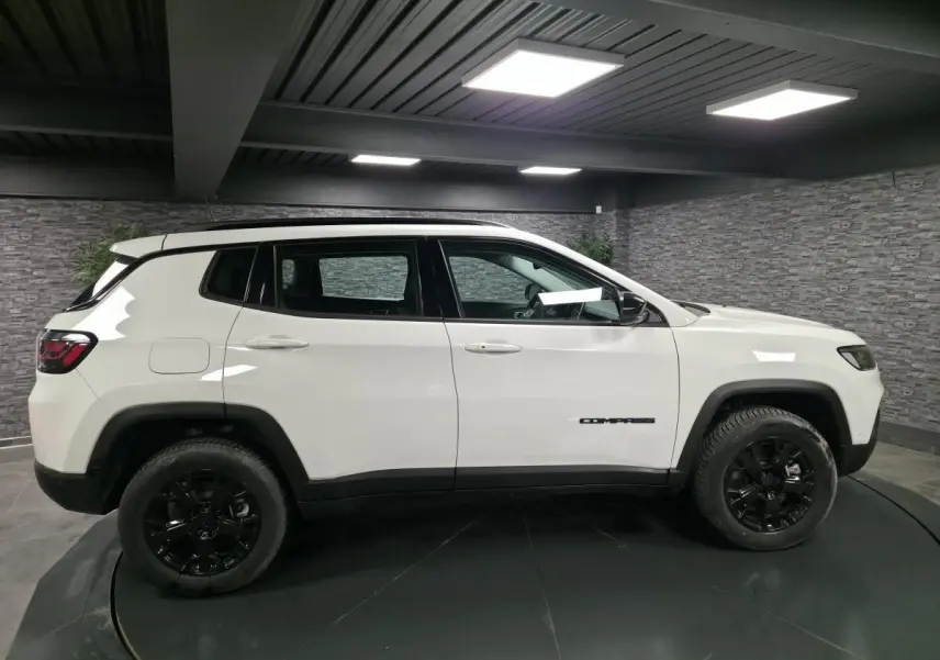 Profil côté gauche d'un Jeep Compass blanc avec jantes noires dans un garage moderne à lumière artificielle.