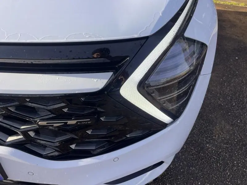 Gros plan sur la calandre noire brillante et le phare avant droit avec éclairage LED en forme de flèche du Kia Sportage blanc nacré GT-Line.