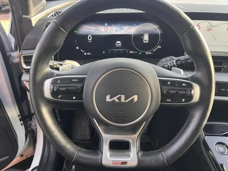 Vue intérieure centrée sur le volant cuir noir GT-line du KIA Sportage blanc, avec tableau de bord digital et écran tactile visible.