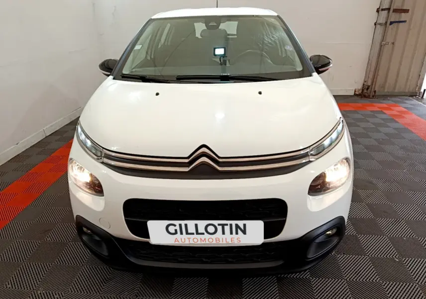 Citroën C3 blanche vue de face en intérieur, avec feux avant allumés et logo distinctif sur la calandre.
