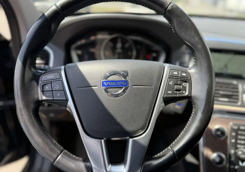 Vue rapprochée du volant en cuir noir du Volvo XC60 gris foncé, avec commandes audio et tableau de bord en arrière-plan.