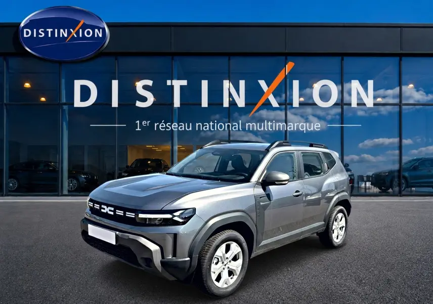 Dacia Duster gris schiste en 3/4 avant droit, avec barres de toit et jantes alu, devant une vitrine Distinxion.