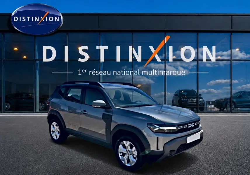 Dacia Duster gris schiste en 3/4 avant droit, avec jantes alu et barres de toit visibles devant vitrine Distinxion.