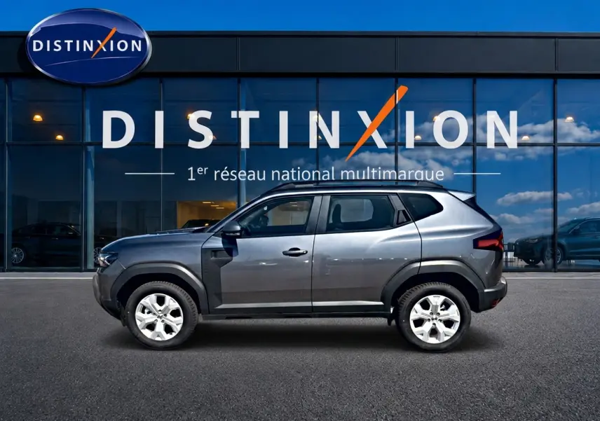 Vue latérale droite d'un Dacia Duster gris schiste 2026 garé devant une vitrine avec logo Distinxion.