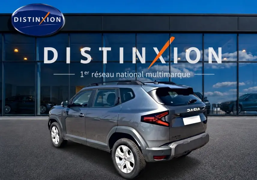 Vue 3/4 arrière droite du Dacia Duster gris schiste 2026 avec barres de toit et jantes alu devant un showroom Distinxion.