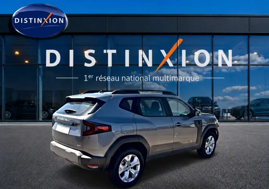 Vue 3/4 arrière droite d'un Dacia Duster gris schiste 2026 avec barres de toit, garé devant une vitrine Distinxion.