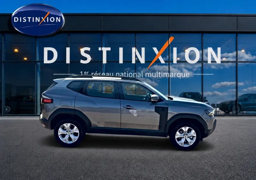 Vue latérale droite du Dacia Duster gris schiste 2026 devant une vitrine Distinxion, soulignant ses jantes alu et barres de toit.