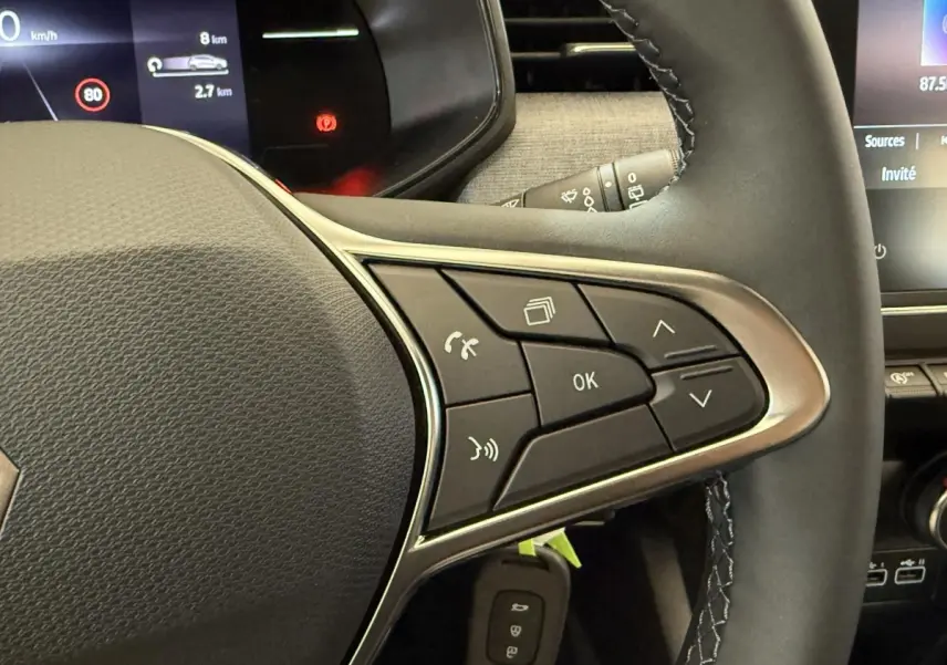 Gros plan sur le volant cuir noir de la Renault Clio 2025, avec commandes multifonctions et tableau de bord digital en arrière-plan.