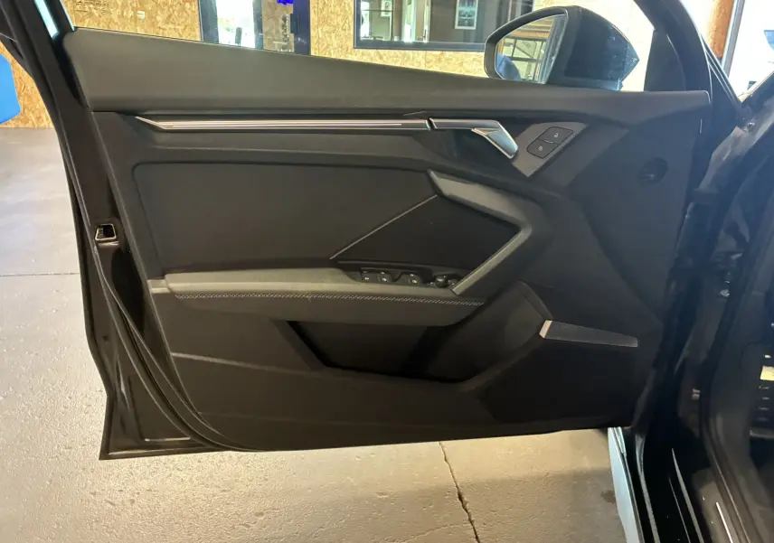 Porte avant gauche ouverte d'une Audi A3 Sportback noire, montrant les commandes de vitres et la poignée intérieure design.