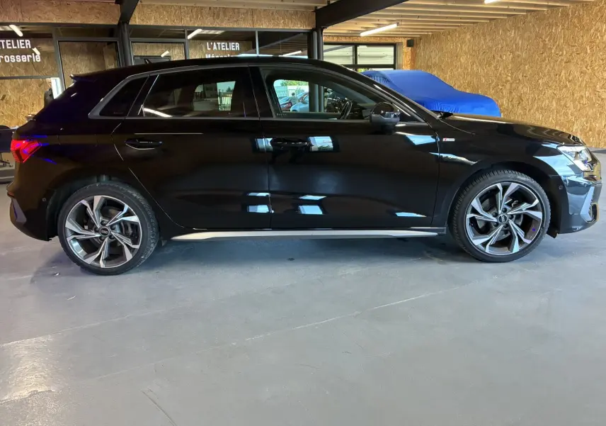 Profil côté gauche d'une Audi A3 Sportback noire 2022 S Line avec jantes alliage et intérieur cuir noir visible en atelier.