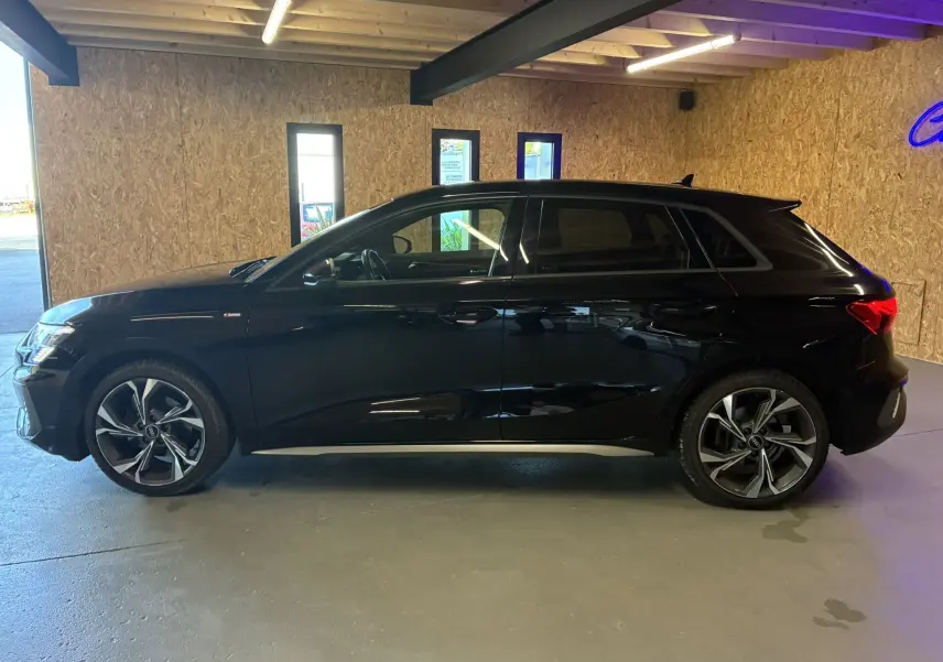 Profil côté gauche d'une Audi A3 Sportback noire 2022 S Line avec jantes alliage et vitres teintées dans un garage.