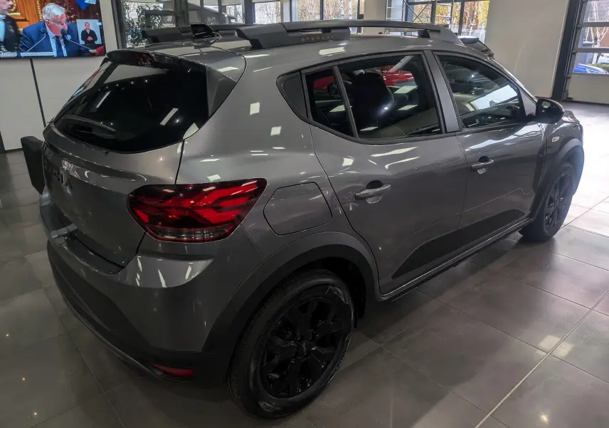 Vue 3/4 arrière droite d'une Dacia Sandero Stepway TCe 110 gris schiste avec jantes noires et barres de toit en intérieur showroom.