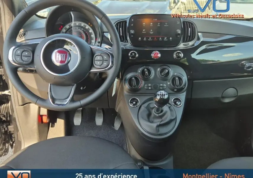 Vue intérieure frontale du tableau de bord noir de la FIAT 500C 2024 avec volant, commandes et levier de vitesse manuel.