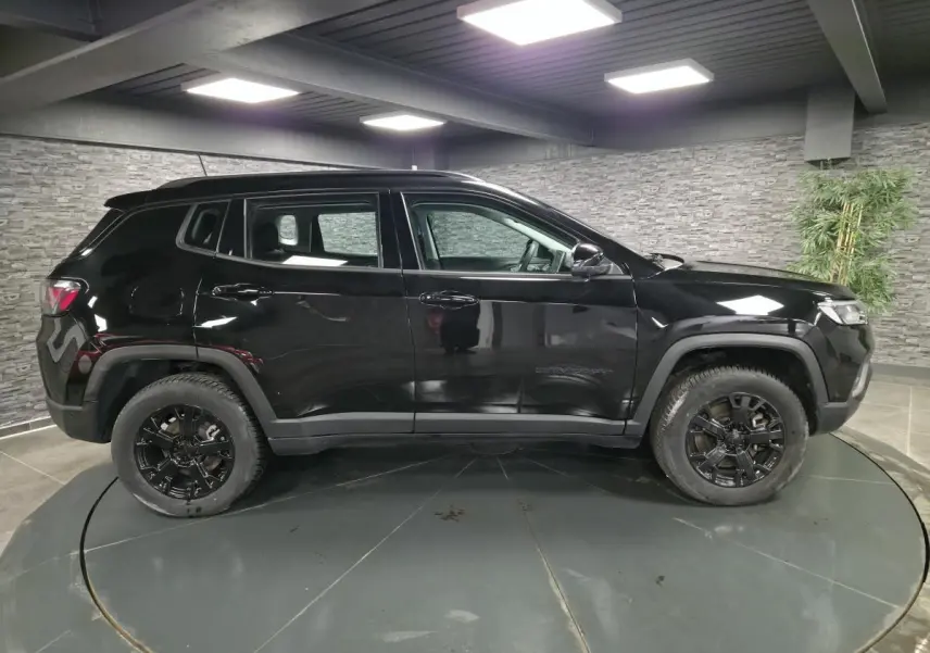 Vue latérale côté gauche d'un Jeep Compass noir 2024 avec jantes noires et garde au sol surélevée dans un showroom.