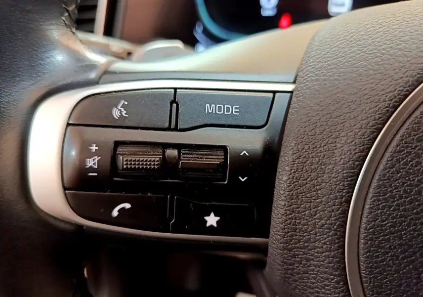 Gros plan sur les commandes gauche du volant cuir noir du Kia Sportage blanc 2022, boutons mode et téléphone visibles.