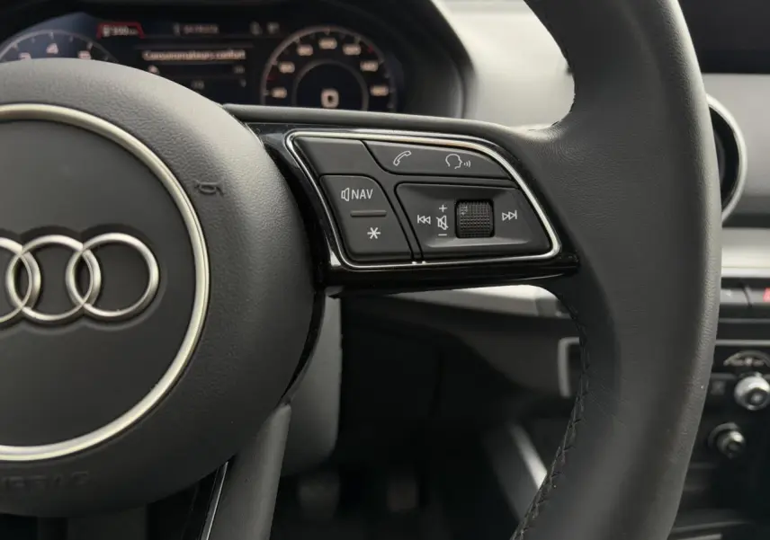 Gros plan sur le volant cuir noir de l'Audi Q2 30 TFSI S-Line 2024 avec commandes multimédia et tableau de bord numérique flou.