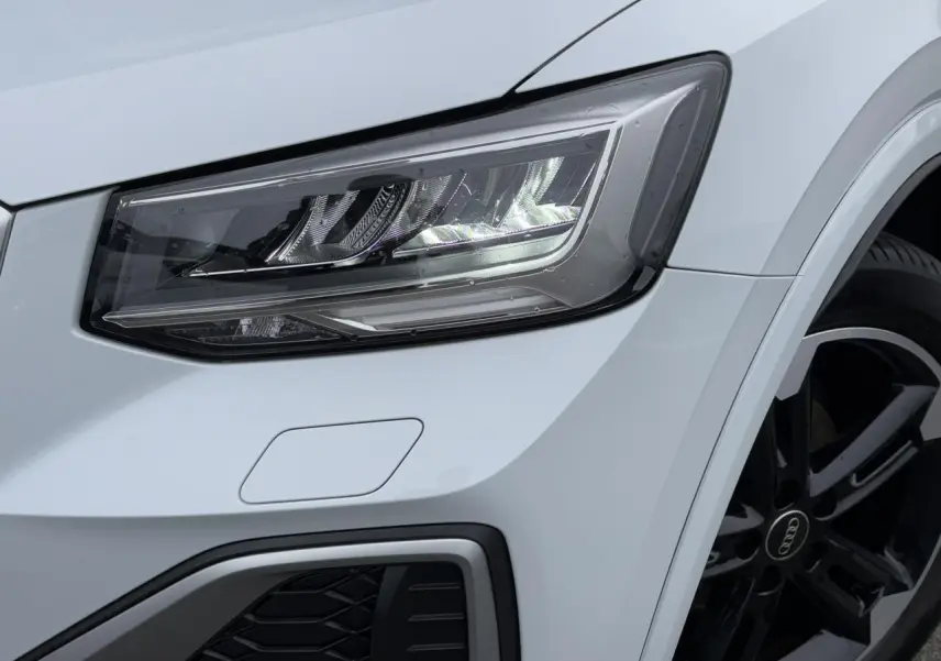 Gros plan sur le phare avant droit et la jante noire polie du SUV Audi Q2 blanc Glacier version S-Line 2024.