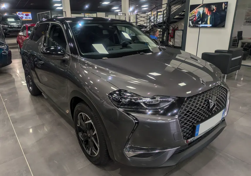 Vue 3/4 avant droit d'un DS3 Crossback gris Platinium avec toit noir et calandre distinctive en losange dans un showroom.