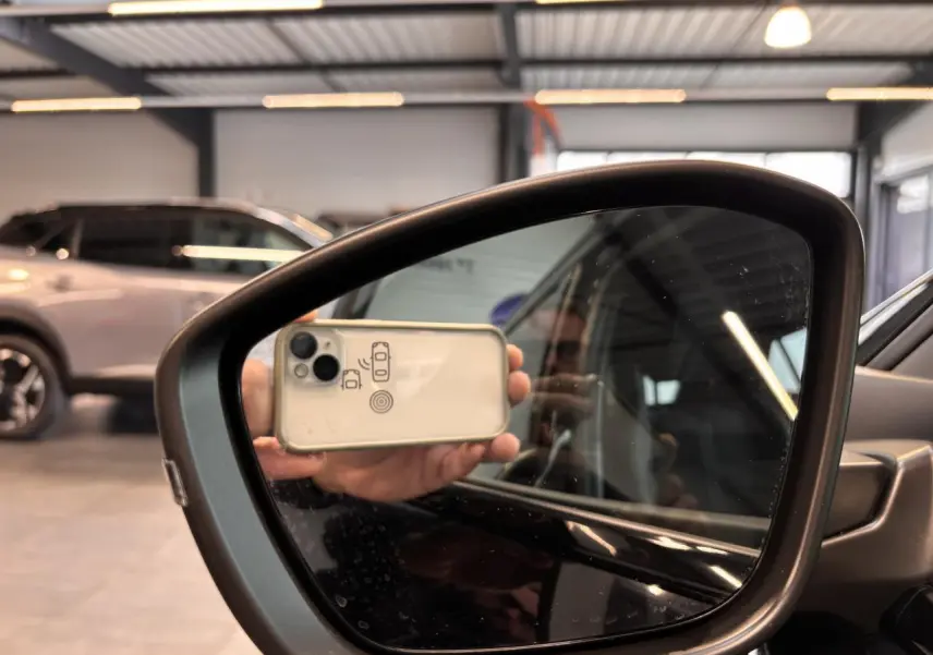 Rétroviseur côté droit du Peugeot 2008 blanc hybride 2025, avec reflet d'un smartphone et intérieur partiel visible.