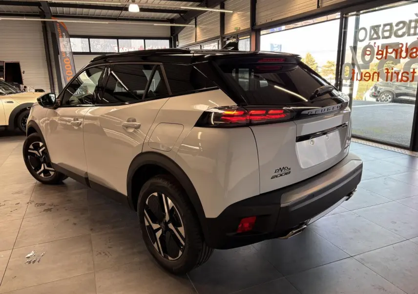 Vue 3/4 arrière droite d'un Peugeot 2008 blanc hybride essence 2025 avec toit noir et feux arrière allumés.