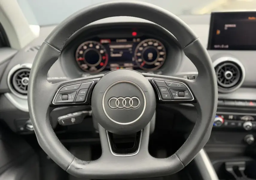 Vue rapprochée du volant cuir multifonction de l'Audi Q2 30 TFSI S-Line 2024 avec tableau de bord numérique en arrière-plan.