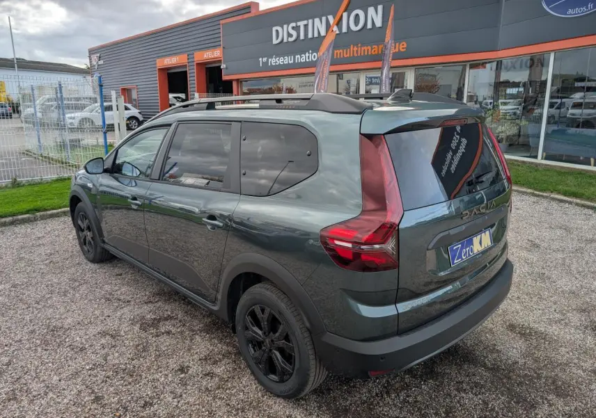 Vue 3/4 arrière droite d'un Dacia Jogger vert métallisé avec jantes noires et barres de toit sur un parking.