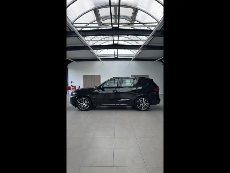 Profil côté gauche d’un BMW X5 xDrive50e noir brillant, avec jantes alliage et toit panoramique en intérieur lumineux.
