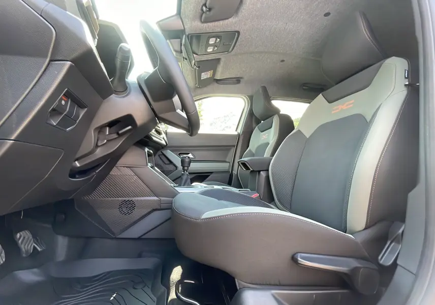 Vue intérieure côté conducteur du Dacia Duster 2025 avec sièges sport gris et logo orange, volant et levier de vitesse manuelle visibles.