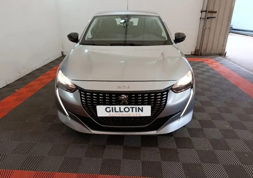 Vue avant d'une Peugeot 208 gris clair avec phares allumés dans un garage fermé.