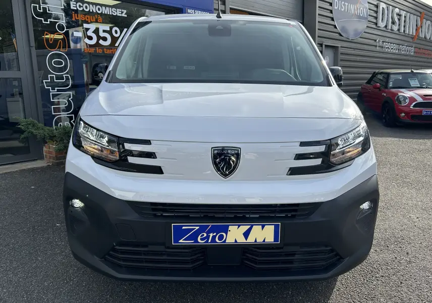 Vue de face d'un Peugeot Partner Fourgon blanc avec logo lion noir et pare-chocs avant noir mat, stationné devant un garage.