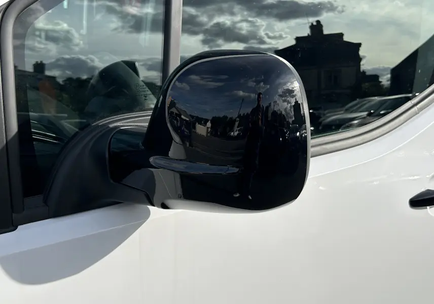 Rétroviseur noir en gros plan côté gauche d'un Peugeot Partner Fourgon blanc, reflet d'un ciel nuageux et d'un bâtiment.