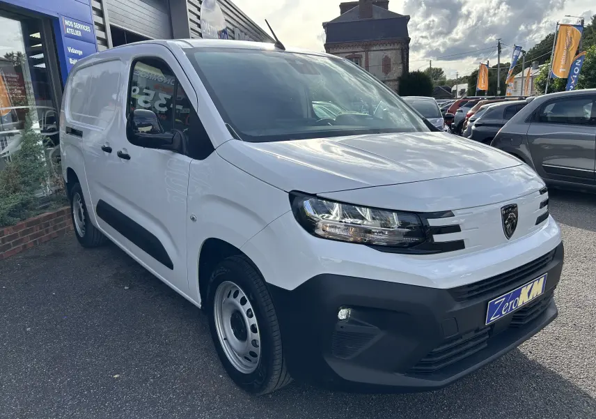 Peugeot Partner Fourgon blanc en 3/4 avant droit avec rétroviseurs noirs et calandre moderne, stationné devant un atelier.