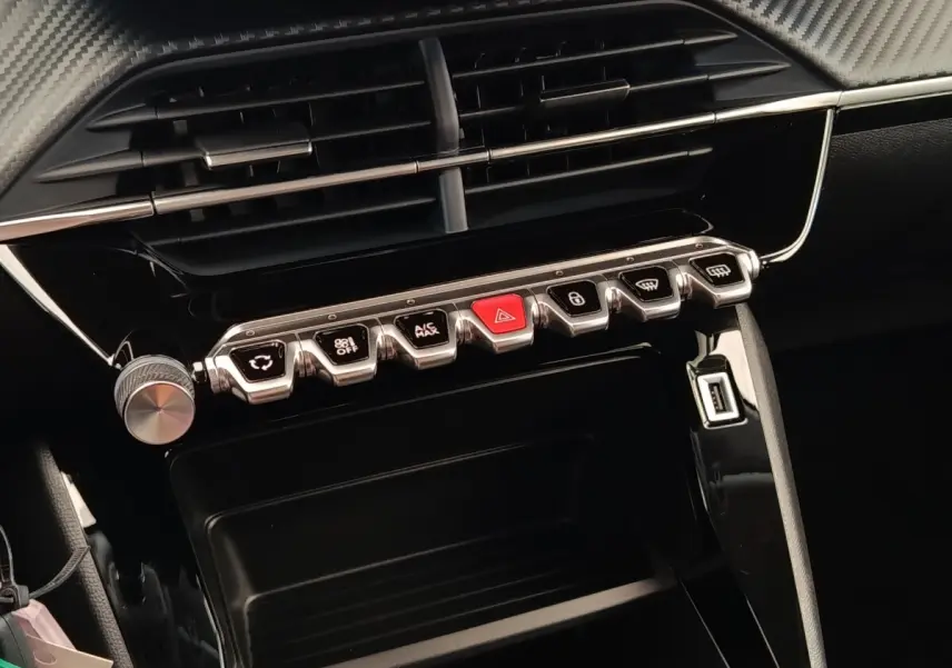 Vue rapprochée du tableau de bord central noir brillant de la Peugeot 208 2023 avec rangée de boutons chromés et bouton rouge d’alerte.