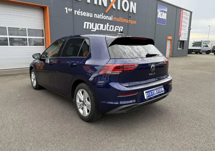 Vue 3/4 arrière droite d'une Volkswagen Golf bleu Atlantic métallisée stationnée devant un bâtiment commercial.