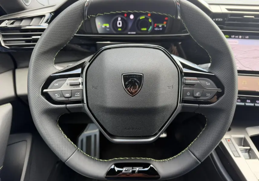 Volant compact GT en cuir noir avec surpiqûres vert Adamite et logo Peugeot, tableau de bord numérique en arrière-plan.