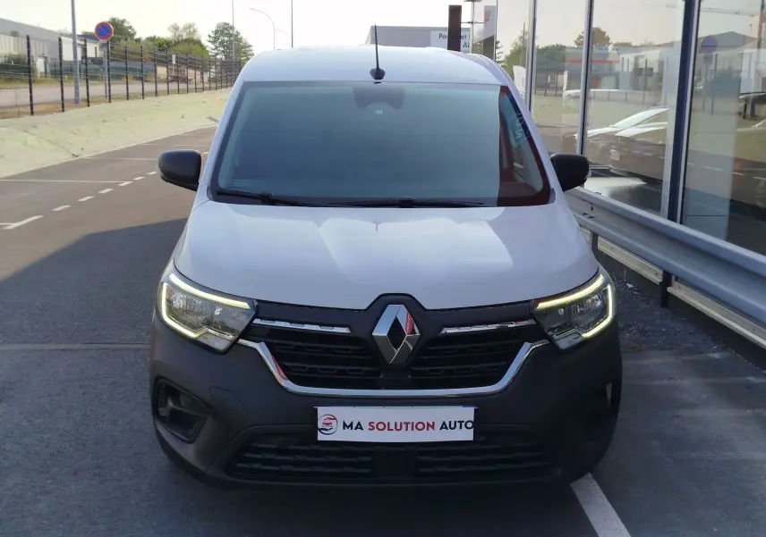 Vue frontale d'un Renault Kangoo blanc minéral 2023 avec calandre noire et feux LED allumés, stationné devant un bâtiment vitré.