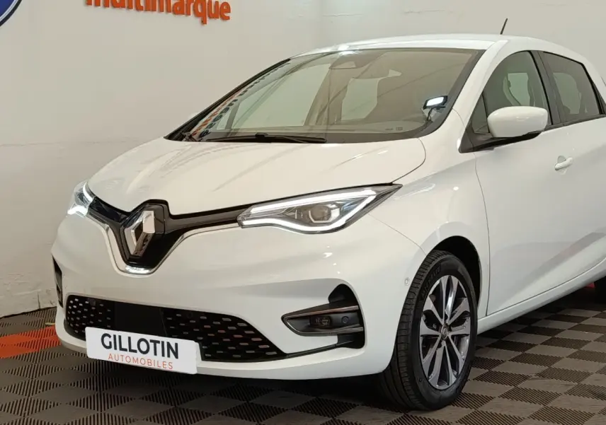 Renault ZOE Intens R110 blanc vue 3/4 avant droit, phares LED et jantes alliage visibles en intérieur showroom.