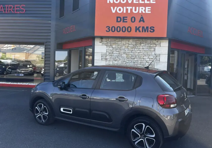 Vue côté gauche d'une Citroën C3 2024 gris foncé avec protections latérales blanches, garée devant un showroom.