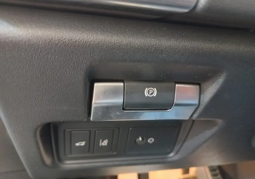 Gros plan sur le bouton de frein à main électrique et commandes du coffre dans l'habitacle noir du Jaguar E-PACE blanc.