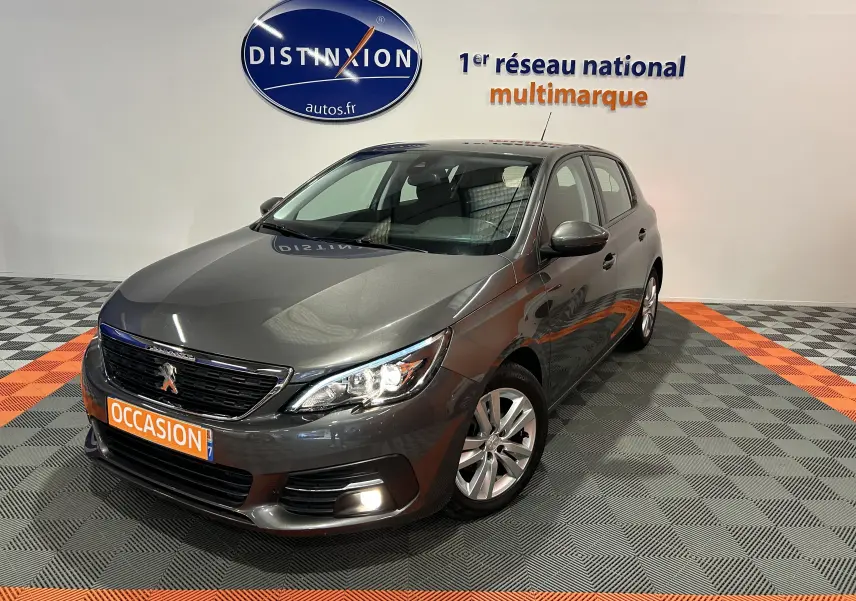 Peugeot 308 gris vue 3/4 avant droit dans showroom avec plaque orange "OCCASION" et logo Distinxion au mur.