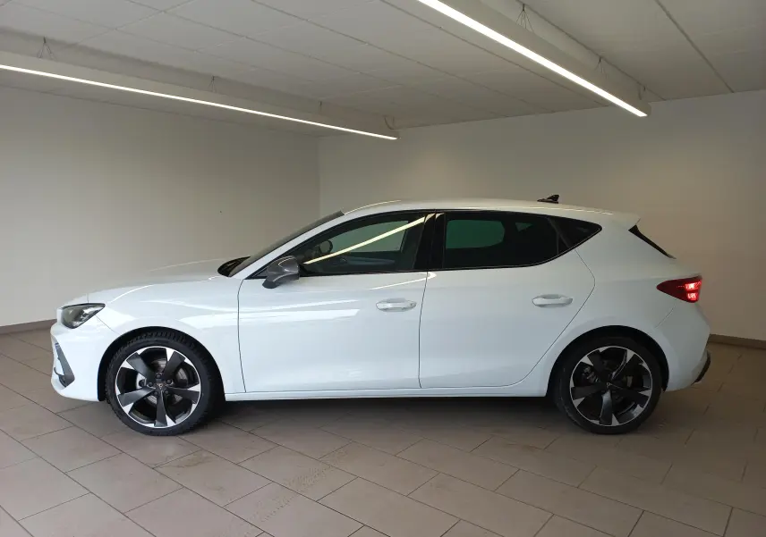 Vue de profil côté gauche d'une CUPRA Leon blanche Nevada avec jantes noires et détails cuivrés, en intérieur.