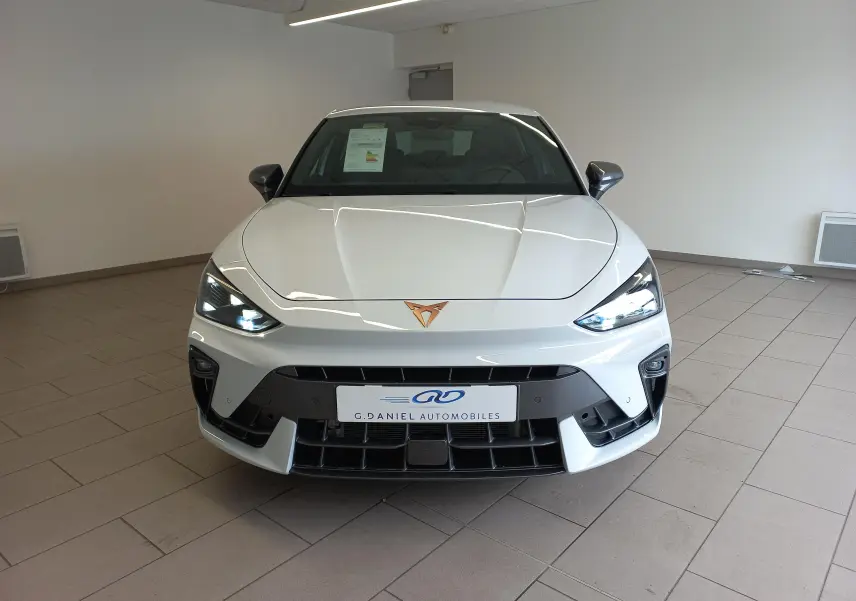 Vue frontale d'une CUPRA Leon Phase 2 blanche Nevada avec feux allumés et logo cuivre visible sur le capot.