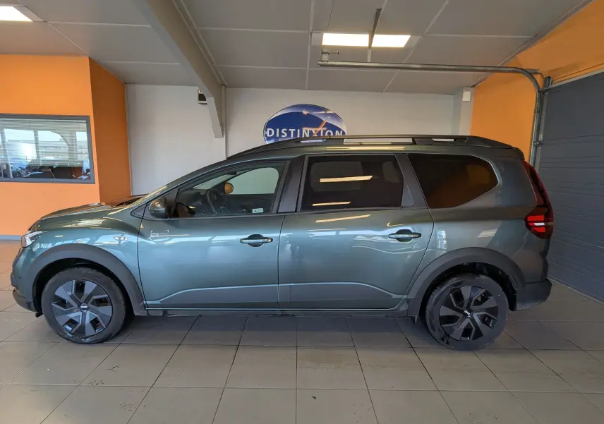 Vue de profil côté gauche d’un Dacia Jogger 2025 vert cèdre métallisé avec jantes noires et toit noir en intérieur showroom.