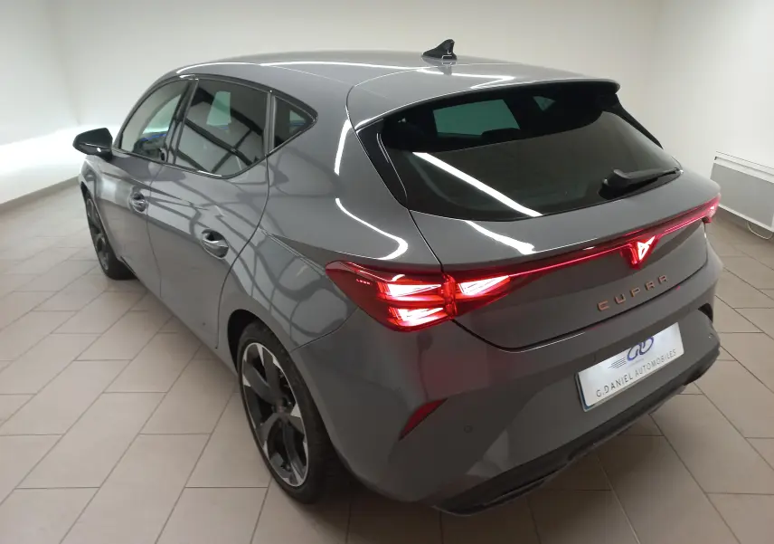Vue 3/4 arrière droite d'une CUPRA Leon Phase 2 gris Graphene avec feux arrière allumés dans un intérieur lumineux.