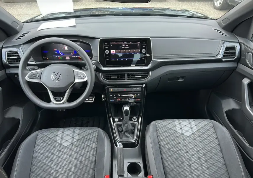 Vue intérieure avant du Volkswagen T-Cross TSI 116 DSG7 R-Line 2025, tableau de bord moderne et sièges tissu gris à motifs.